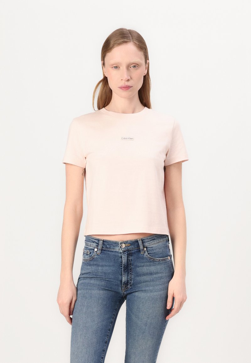 Šviesiai rožinė trumpa medvilninė marškinėliai su apvalia kaklo iškirpte ir mažuoju Calvin Klein logotipu ant krūtinės. Suderinta su mėlynomis džinsinėmis kelnėmis.