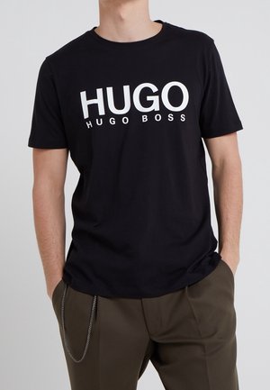 Homme portant un t-shirt noir avec le grand texte blanc "HUGO" et un texte plus petit "HUGO BOSS", assorti à un pantalon olive foncé et un accessoire en chaîne latérale.