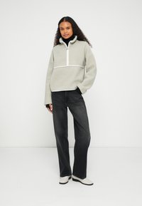 Jasnoszara bluza polarowa z wysokim kołnierzem, zamkiem błyskawicznym i białymi akcentami, zestawiona z czarnymi jeansami o szerokich nogawkach i kremowymi mokasynami.