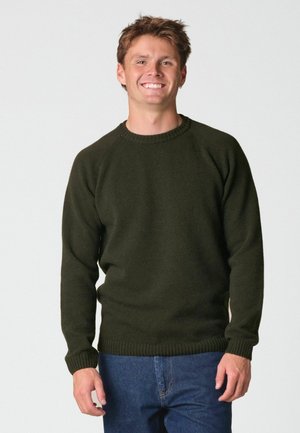 Jeune homme souriant, portant un pull en maille vert foncé et un jean bleu, debout devant un fond clair uni.