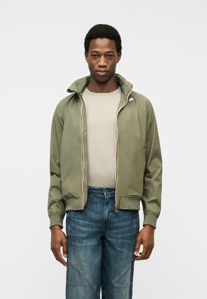 Jauns vīrietis, kurš valkā olīvu zaļu rāvējslēdzēja jaku, bēša krāsas kreklu un zilas džinsas, stāv pret vienkāršu gaišu fonu.