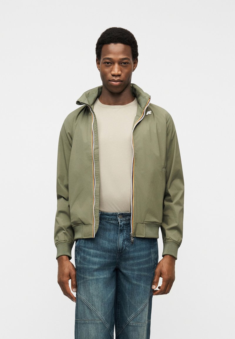 Νεαρός άνδρας που φοράει μπουφάν χακί με φερμουάρ, μπεζ πουκάμισο και μπλε τζιν, στέκεται μπροστά σε απλό φωτεινό φόντο.