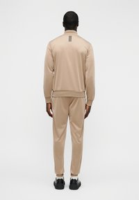 Μπεζ φόρμα από λείο ύφασμα, με ψηλό γιακά και οριζόντια ρίγα στο πίσω μέρος. Φορεμένη με μαύρα παπούτσια.