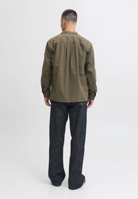 Camisa de manga larga color verde oliva con corte recto y detalles de costura visibles, combinada con pantalones de mezclilla anchos en un tono oscuro.