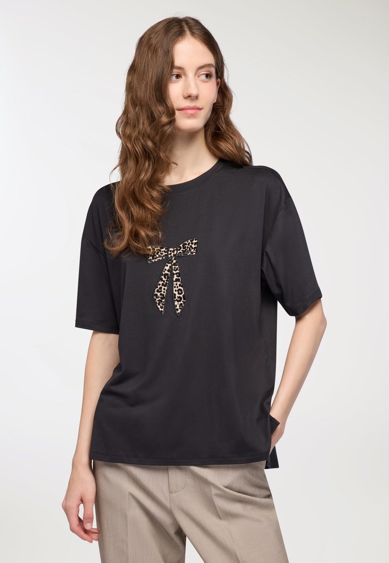 T-shirt nera a maniche corte con un dettaglio a fiocco leopardato sulla parte anteriore. Tessuto morbido, vestibilità rilassata, abbinata a pantaloni color marrone chiaro.