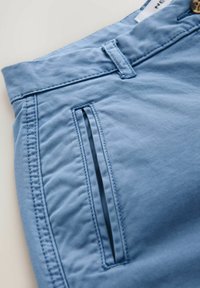Pantalon bleu clair avec une poche fente verticale, passant de ceinture et coutures visibles sur la ceinture et la couture latérale.