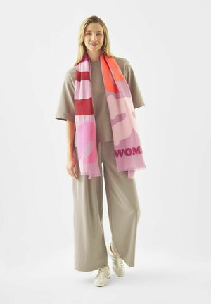 Vrouw draagt een beige loszittende outfit en witte sneakers, gecombineerd met een lange sjaal in roze, rood en oranje patroon.