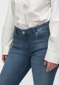 Persona che indossa jeans di denim blu e camicia bianca con bottoni, con le mani rilassate lungo i fianchi.