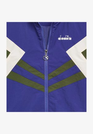 Blauwe jas met ritssluiting aan de voorkant, witte en groene chevronpatronen op mouwen en borst, en het "diadora"-logo aan de rechterbovenkant.