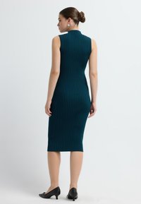 Abito a maglia a coste verde acqua con colletto alto e design senza maniche, che arriva a metà polpaccio. Abbinato a scarpe nere con tacco basso.