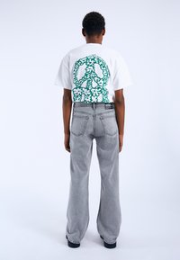 Camiseta blanca con signo de paz verde y un patrón de flores en la parte posterior, combinada con jeans de campana grises de cintura alta. Footwear negro visible.