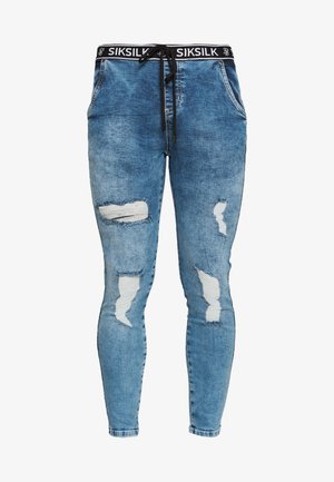 Joggers en denim bleu clair avec une ceinture élastique noire portant le nom de la marque. Délavés avec des déchirures et des détails effilochés sur les jambes.