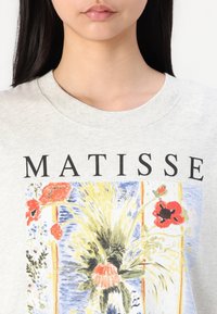 Pilkas džemperis su spalvingu Matisse stiliaus printu, kuriame pavaizduoti gėlių raštai raudona, geltona ir mėlyna spalvomis. Drąsiais juodais rašmenimis užrašyta "MATISSE."