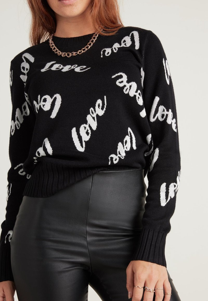 Personne portant un pull noir avec un motif texte blanc "love", un pantalon en cuir noir, un collier chaîne et des bagues aux doigts, debout devant un fond uni.