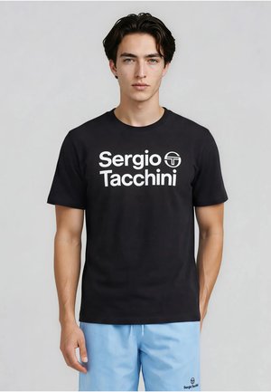 Ung mand med mørkt hår iført en sort Sergio Tacchini t-shirt og lyseblå shorts, der står foran en ensfarvet grå baggrund.