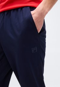 Άτομο που φοράει σκούρο μπλε παντελόνι με το χέρι στην τσέπη, με ένα μικρό μαύρο λογότυπο, και κόκκινο πουκάμισο που φαίνεται μερικώς.