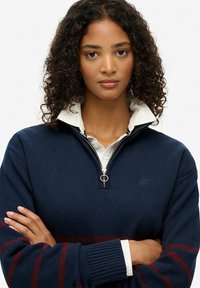 Granatowy sweter typu pullover z ćwierć zamkiem, z białą koszulą pod spodem. Bordowe poziome paski na rękawach, teksturowany materiał.