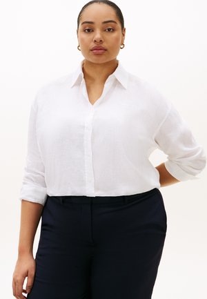 Woman wearing a white button-up shirt with rolled-up sleeves and black pants, standing with one hand behind her back.
