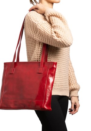 Vrouw in beige gebreide trui draagt grote rode leren shopper aan de schouder tegen een witte achtergrond.