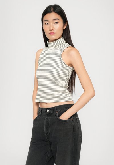 Junge Frau mit langen dunklen Haaren, die einen ärmellosen, beigen, gestreiften Rollkragenpullover und schwarze, hochtaillierte Jeans trägt, mit den Händen in den Taschen.