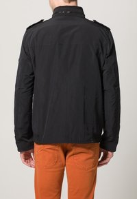 Czarna lekka kurtka o gładkiej fakturze, z pagonami na ramionach i prostym krojem, zestawiona z pomarańczowymi dżinsami.