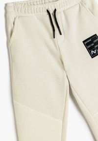 Lys beige sweatpants med ribbet tekstur, elastisk talje, sorte snore, sidelommer og en lille sort patch-detalje.