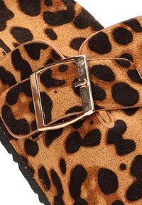 Sandal de estampado de leopardo con una textura similar al ante, diseño plano y un acento de hebilla de metal dorado. Manchas oscuras sobre un fondo marrón claro.