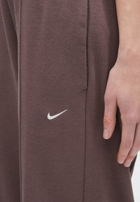 Close-up de uma mão repousando ao lado de calças Nike castanhas com um logo swoosh branco perto da costura do bolso lateral.