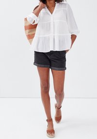 Blouse blanche texturée avec fermeture à boutons sur le devant et design à étages ; associée à un short en denim noir retroussé et des sandales marron à lanières.