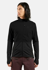 Schwarze Zip-Jacke aus einem glatten, dehnbaren Material. Mit hohem Kragen, Seitentaschen und Daumenöffnungen an den Manschetten.