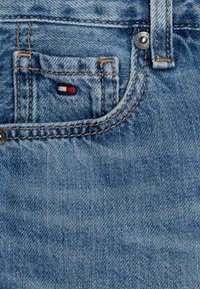 Jasnoniebieska tkanina dżinsowa z małą naszywką logo w kolorze czerwonym, białym i niebieskim na tylnej kieszeni. Posiada widoczne szwy i metalowy nit.