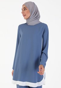 Tunic blu con maniche lunghe, scollo rotondo e dettagli bianchi all'orlo. Tessuto morbido con una texture liscia, completato da un hijab grigio chiaro.