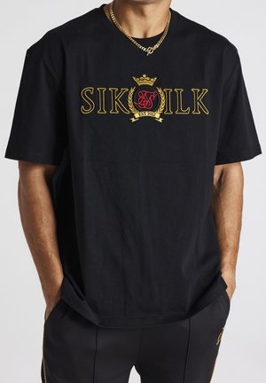 Man draagt een zwart SikSilk t-shirt met goud geborduurd logo en wapen, gecombineerd met zwarte broek, handen in de zakken en een gouden ketting.