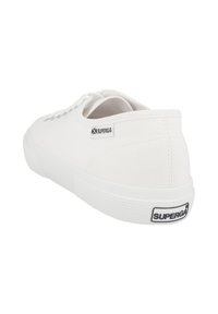 Sneaker bianca in tela con punta rotonda, suola piatta e design con lacci. Presenta un'etichetta con logo sul lato e suola in gomma testurizzata.