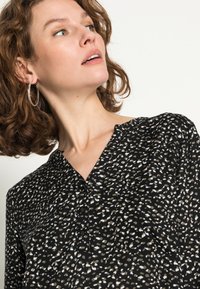 Blusa preta com padrão, apresentando um design salpicado de branco e castanho, com decote em V e detalhes em botão na frente. Tecido macio.