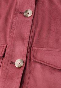 Pink ruskindsjakke tæt på med cremefarvede knapper og to rektangulære klaplommer foran.