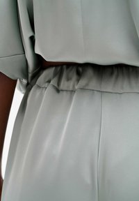 Lichtgrijze top van gladde stof; heeft korte mouwen, een elastische tailleband en zachte plooien voor extra textuur en flow.