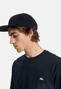 Gorra negra con visera plana y logo, combinada con una camisa de color azul oscuro, de manga corta y texturizada, que presenta un pequeño detalle de logo blanco.