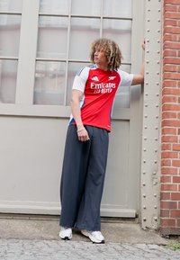 Tricou de fotbal Adidas roșu și alb cu logo-ul "Emirates", purtat cu pantaloni lărgi gri închis și adidași albi, stând în fața unui perete.