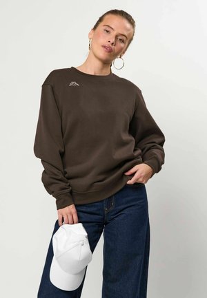 Femme portant un sweat-shirt marron foncé et un jean bleu, tenant une casquette de baseball blanche, debout devant un fond uni clair.