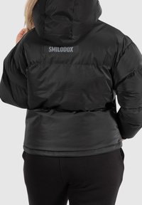 Giacca imbottita nera con cappuccio, caratterizzata da una texture liscia e lucida. Sul retro è presente un logo "SMILODOX" su una toppa rettangolare.