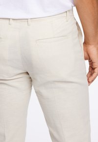 Zbliżenie na osobę w jasnobeżowych lnianych spodniach i białej koszuli, ukazujące tylne kieszenie i część prawego ramienia.