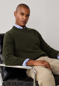 Dunkelgrüner Pullover mit rundem Ausschnitt und gerippten Bündchen, über einem hellblau gestreiften Hemd getragen, kombiniert mit beigen Hosen.