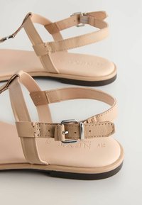 Beige leather sandals with slim straps, adjustable ankle buckle, flat soles, and subtle stitching details. Low-profile design.
