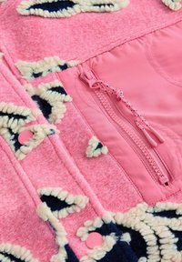 Rožnata flis tkanina z reliefnim vzorcem, poudarjenim s kremnimi poudarki in mornarsko modrimi obrobi. Vključuje rožnati zadrgo in pritiskače za zapiranje.