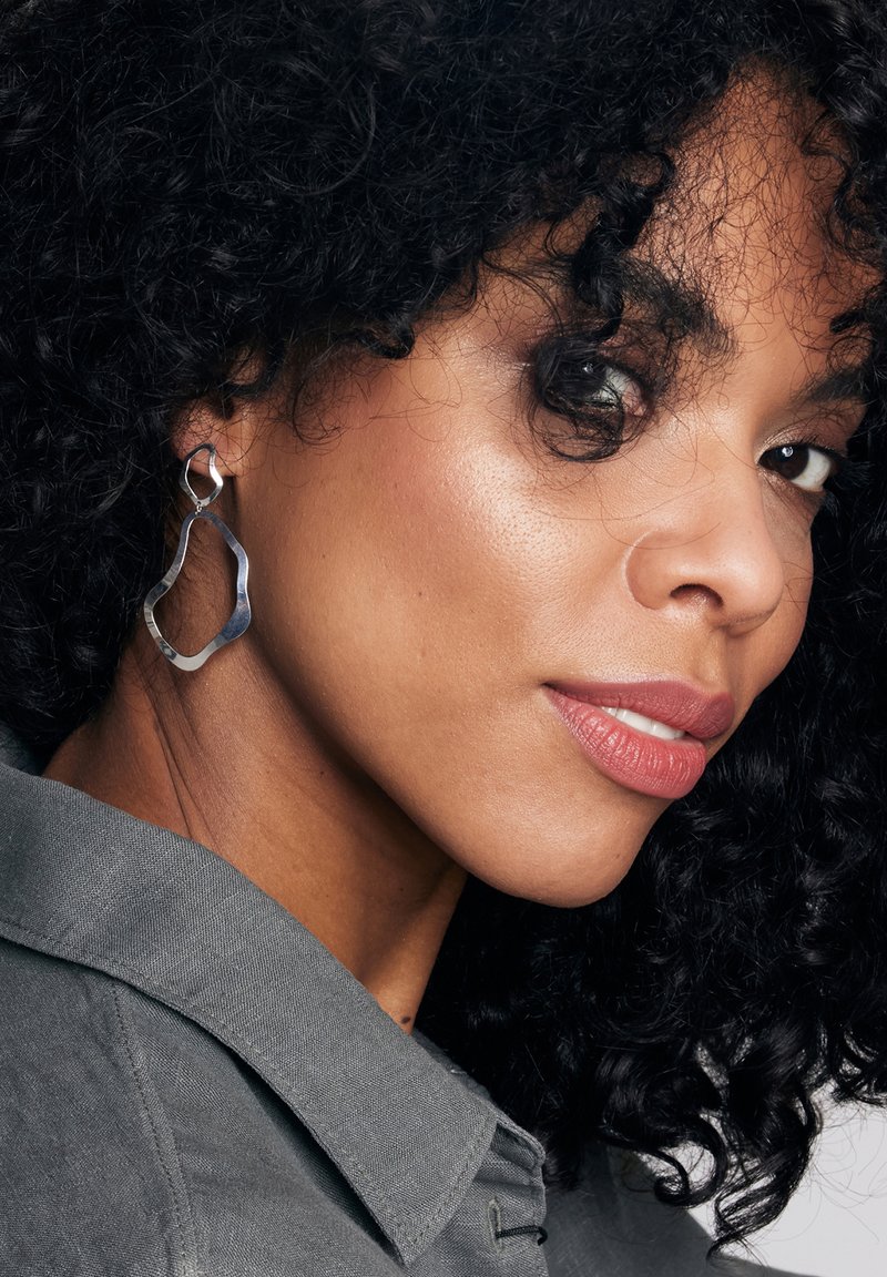 Srebrne metalowe kolczyki w abstrakcyjnym kształcie o gładkiej, polerowanej powierzchni. Prezentowane na osobie z kręconymi włosami, noszącej szary T-shirt.