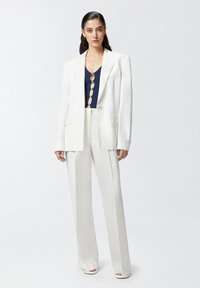 Bijela krojena blazerica i odgovarajuće široke hlače uparené s mornarsko plavom majicom s detaljem metalne dugmadi. Glatka tekstura tkanine.