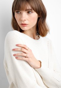 Maglione bianco lavorato a maglia con una trama larga, che mostra una superficie a coste. La modella indossa anelli sulle dita, evidenziando delicati dettagli in oro e argento.