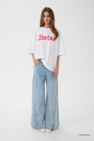 Woman standing wearing a white oversized "Barbie" t-shirt, wide-leg light blue jeans with buttons, and white pointed shoes against a plain background.