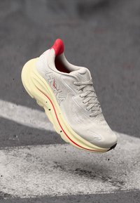 Scarpe da corsa Hoka bianche e rosse sospese in aria sopra l'asfalto con piccoli detriti e segnaletica stradale bianca.